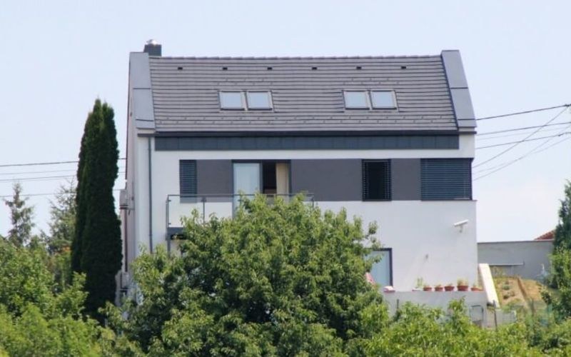 A ház a kert felől: nyeregtető tetőtéri ablakokkal, erkély, előtte lombos fák.